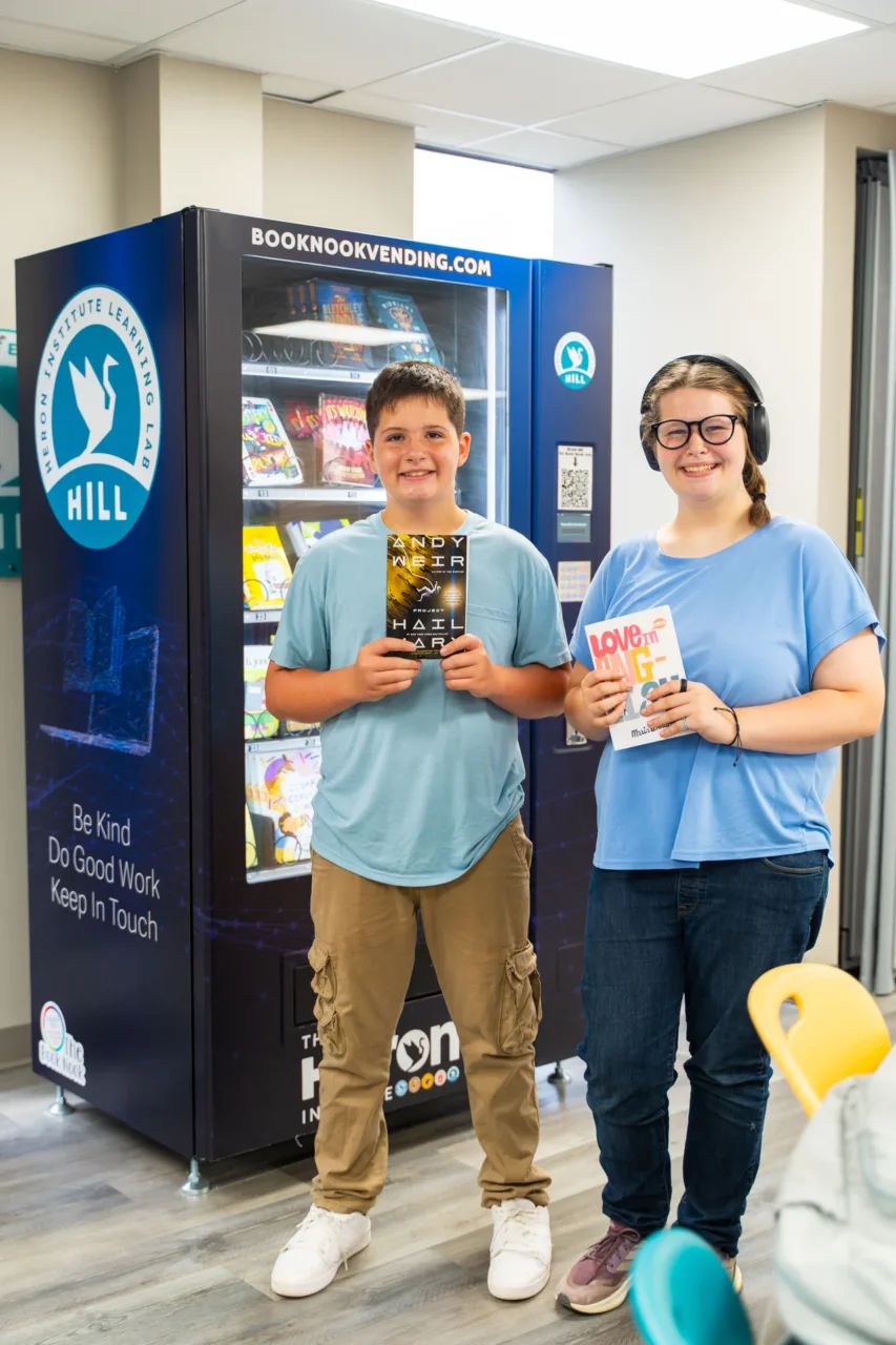 Two students at Heron Institute Learning Lab smile, holding books. A vending machine promotes positivity.