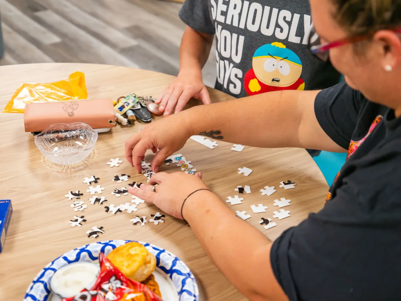 Hands assembling a puzzle at a wooden table with snacks nearby, symbolizing The HILL's community support.