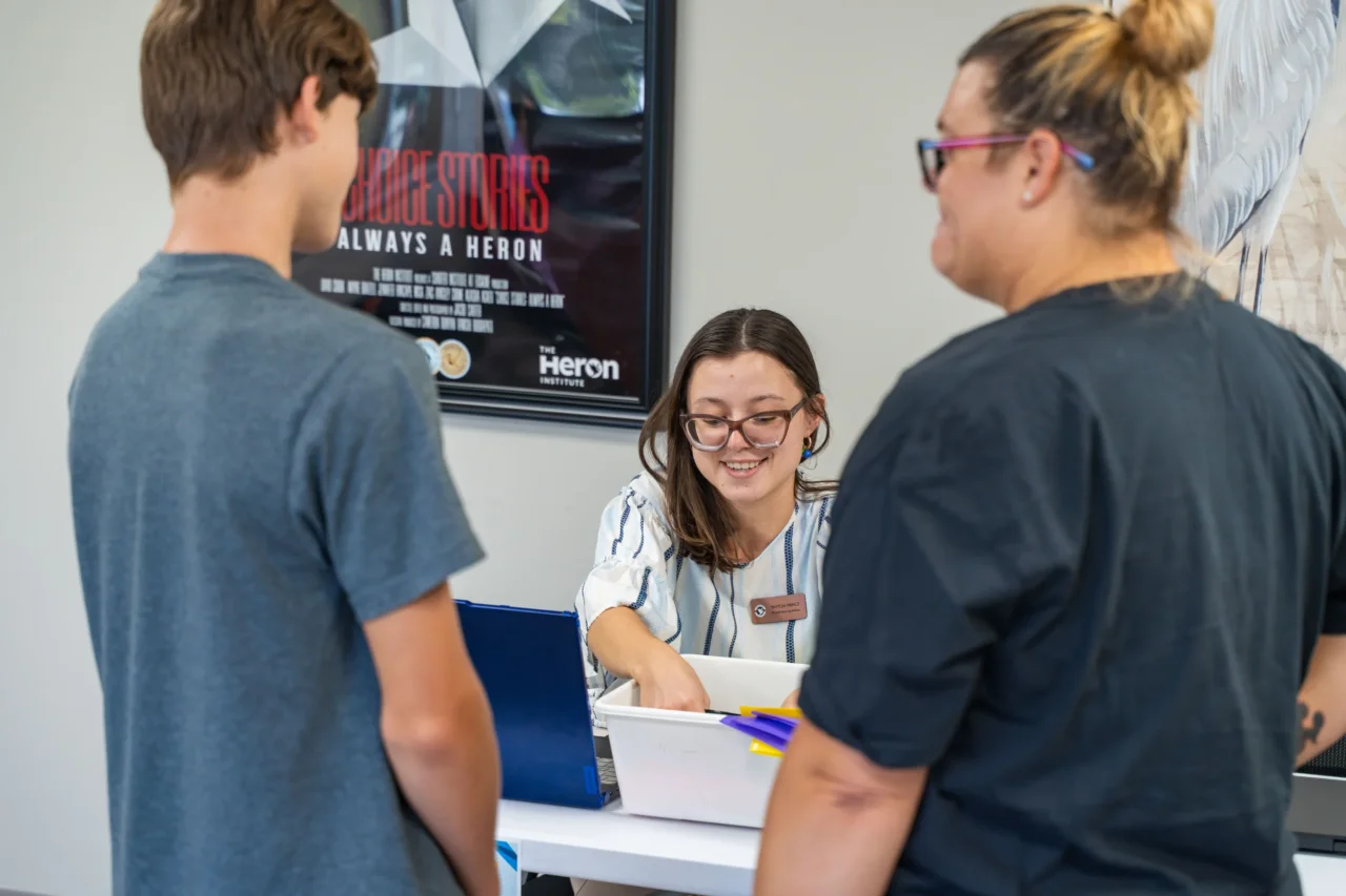 A smiling staff member assists two people, highlighting The HILL's support for students enrolled in Cyber or Heron Virtual Academy.