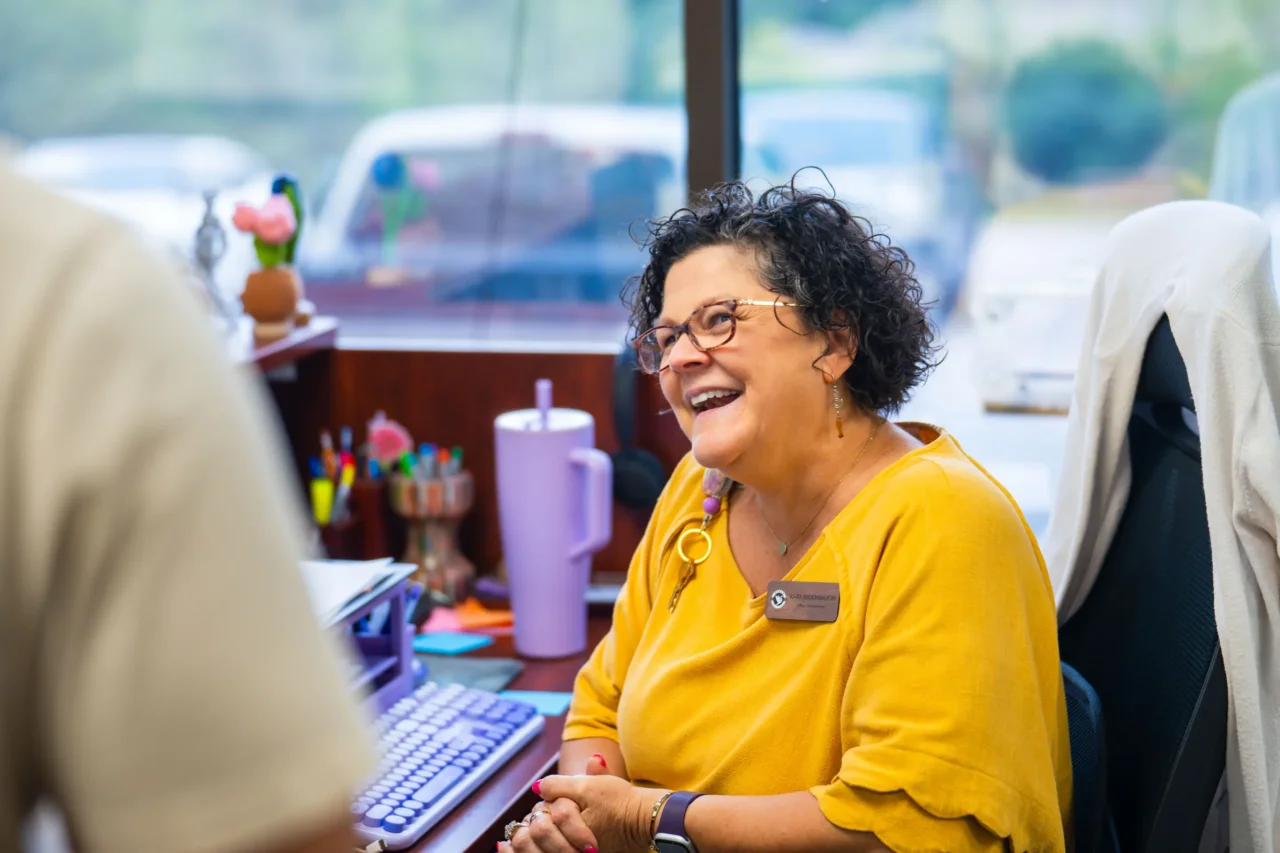 Smiling woman in a yellow top at a desk in a cyber academy, fostering a welcoming school community.