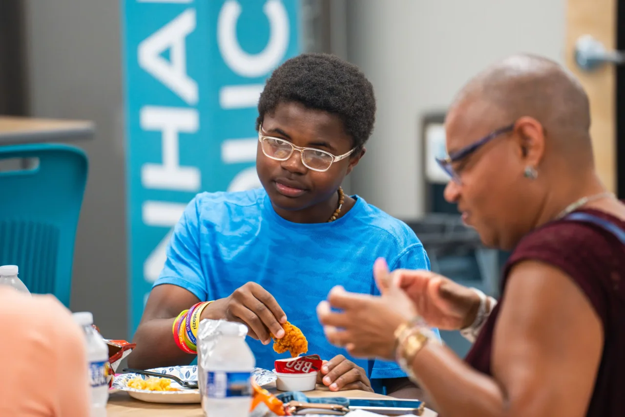 Person in a blue shirt enjoying food at a community amp event, engaging in conversation with another attendee.