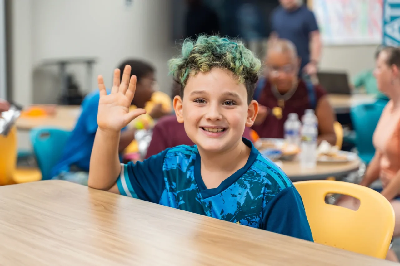 Smiling child with raised hand sitting in a vibrant, shared learning space, engaging with peers.